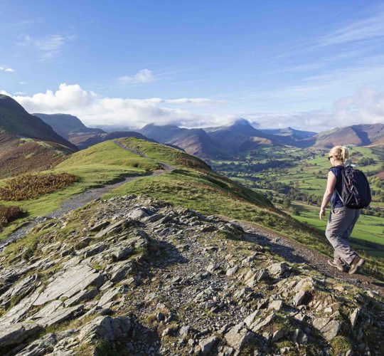 Woman hiking on a rocky hilltop path overlooking rolling green valleys and distant peaks in the Lake District on a clear, sunny day. ?? ''